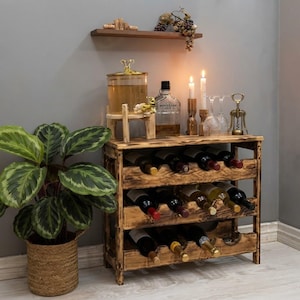 Puede incluir: Un botellero de madera rústica con tres estantes que contiene varias botellas de vino. Encima hay un dispensador de bebidas de cristal, una botella de licor, dos velas encendidas, dos copas de vino y un sacacorchos de metal. Una planta al lado.