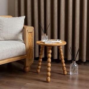 May include: A light wood side table with a round top and three legs featuring a stacked ball design. On the table sits a glass reed diffuser and two stacked books. A second glass reed diffuser with black reeds stands on the floor nearby.
