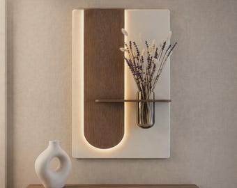 May include: A wall-mounted rectangular panel featuring a white base and a dark wood vertical accent with integrated backlighting. A small horizontal shelf holds a glass vase containing dried lavender and white fluffy botanical stems.