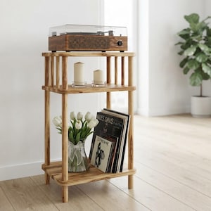 May include: A three-tiered wooden stand with a light brown finish. The top shelf holds a wood-grain record player with a clear lid. The middle shelf features two white pillar candles. The bottom shelf contains a glass vase with white tulips and vinyl records.