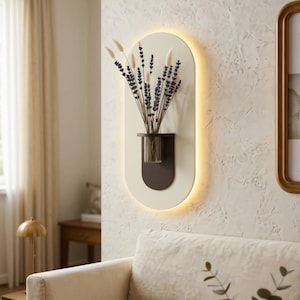 Könnte beinhalten: Eine ovale Wandleuchte mit einer cremefarbenen Rückplatte und einem dunkelbraunen Regal, das eine klare Glasvase hält. Die Vase enthält einen Strauß aus getrocknetem Lavendel und weißen Grashalmen. Die Leuchte strahlt ein sanftes Licht aus.