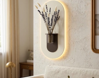 May include: An oval wall-mounted light fixture with a cream-colored backplate and a dark brown shelf holding a clear glass vase. The vase contains a bouquet of dried lavender and white grass stems. The fixture emits a soft glow from behind.