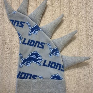 May include: A gray fleece hat featuring a repeating blue Detroit Lions logo and text pattern. The hat has a wide gray brim and five pointed, cone-shaped gray fleece spikes extending from the top edge, resembling a mohawk or dragon crest.