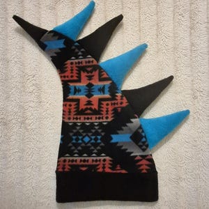 May include: A fleece hat featuring a black base and a geometric pattern in shades of orange, blue, gray, and black. The hat includes a series of alternating black and bright blue pointed spikes extending from the top and back of the headpiece.