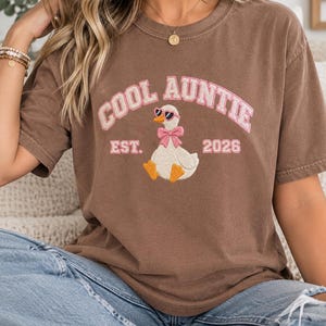 Puede incluir: Una camiseta marrón de manga corta con un bordado de un pato blanco que lleva gafas de sol rosas en forma de corazón y un lazo rosa. El texto COOL AUNTIE EST. 2026 aparece en letras rosas con un borde blanco sobre y bajo el pato.