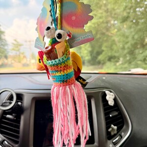 May include: A crocheted holder with a multi-colored striped pattern, two large white googly eyes, and long pink fringe tassels. The holder contains two small tubes and hangs from a rearview mirror alongside a tie-dye Cotton Candy air freshener.
