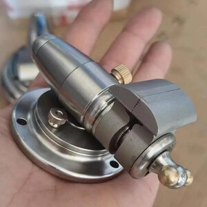 May include: A small silver-toned metal bench vise featuring a circular base with mounting holes, a cylindrical body, and a textured brass adjustment knob. The vise includes a horizontal clamping mechanism with a brass ball-ended handle for tightening.