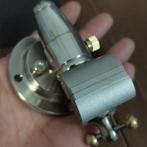 May include: A silver-toned metal bench vise featuring a circular mounting base with pre-drilled holes, a cylindrical body, and a rectangular clamping jaw. The tool includes small brass-colored adjustment knobs and a polished metallic finish throughout.