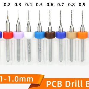 May include: A set of ten PCB drill bits ranging from 0.1 to 1.0 mm in diameter. Each silver metal bit is held in a clear plastic case with a color-coded ring at the base. The sizes 0.1, 0.2, 0.3, 0.4, 0.5, 0.6, 0.7, 0.8, 0.9, and 1.0 are labeled above.
