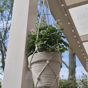 Op de afbeelding: Een hangende plantenbak met een crèmekleurig macramé net van touw rondom een bijpassende crèmekleurige pot. De pot bevat een groene plant met kleine roze bloemetjes. Het geheel hangt aan een dik, geknoopt touwkoord.