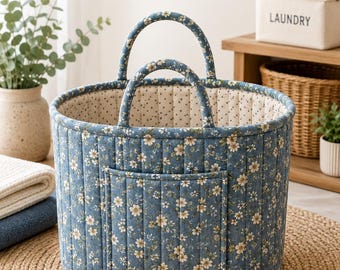 May include: A blue quilted laundry bucket bag sewing pattern featuring a floral print, two padded handles, and a front storage pocket. The text reads LAUNDRY BUCKET BAG sewing pattern, BEGINNER FRIENDLY, A0 A4 US LETTER, 2 SIZES REGULAR & LARGE, FRONT POCKET FOR STORAGE, QUILTED & STRUCTURED, PADDED HANDLES.