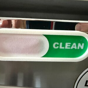 May include: A white plastic dishwasher magnet slider with a rectangular frame. The slider features a green panel with the white text CLEAN visible on the right side, while the left side shows a solid white panel covering the status indicator.