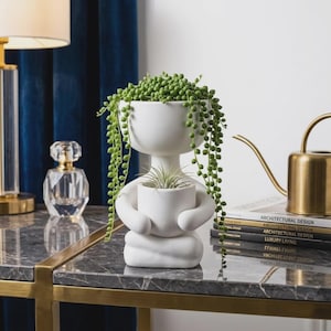 May include: A white ceramic planter shaped like a person sitting in a cross-legged position, holding a smaller white pot. The larger pot contains a trailing green string of pearls plant, while the smaller pot holds a spiky air plant.
