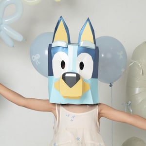 Puede incluir: Una máscara de papel con la forma del personaje Bluey, en tonos azul, blanco y negro. La máscara se lleva con un vestido sin mangas de color crema con pequeños motivos florales azules, calcetines de canalé beige y bailarinas claras.