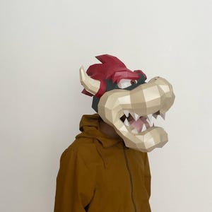 Könnte beinhalten: Eine Low-Poly-Papiermaske der Figur Bowser mit roter Mähne, cremefarbener Schnauze, spitzen weißen Zähnen und geschwungenen Hörnern. Die Maske wird über dem Kopf getragen und mit einer senfgelben Kapuzenjacke mit Reißverschluss kombiniert.