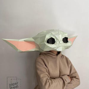 Puede incluir: Una máscara de papel low-poly de una criatura con orejas grandes y puntiagudas, piel verde claro y ojos negros. La máscara presenta detalles rosados en el interior de las orejas y un diseño geométrico facetado. El texto Paper Shapes es visible en la máscara.
