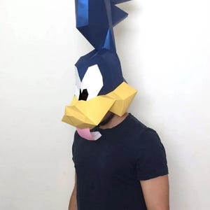 Puede incluir: Una máscara de papel de estilo low-poly con forma de cabeza de pájaro de dibujos animados. La máscara presenta una cresta alta de color azul oscuro, un pico amarillo, parches oculares blancos con pupilas negras y una pequeña lengua rosa.