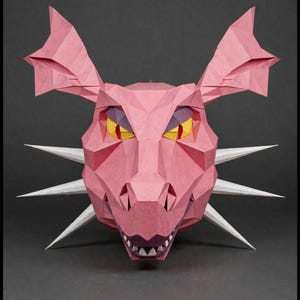 May include: A low-poly papercraft dragon head sculpture in shades of pink. The creature features large pointed ears, sharp white spikes protruding from the sides, and yellow eyes with red pupils. The mouth is slightly open, revealing white teeth.