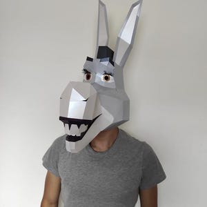 Puede incluir: Una máscara de papel low-poly de una cabeza de burro con una superficie geométrica gris claro, orejas altas y puntiagudas, ojos marrones con largas pestañas negras y una boca abierta que muestra dientes blancos, usada sobre la cabeza.