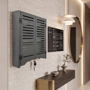 May include: A dark gray wall-mounted key cabinet featuring two louvered doors with ornate metal hinges. The cabinet has a cutout bottom edge and a set of keys hanging from a hook. The rectangular unit is mounted on a light-colored stone wall.