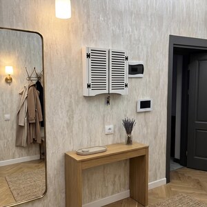 May include: A white wall-mounted key cabinet with louvered doors, a light oak console table, a tall rectangular mirror with a gold frame, and a glass vase containing dried lavender, all arranged in a hallway with light textured wallpaper.