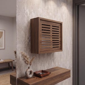 Può includere: Un mobiletto rettangolare da parete in legno con pannello frontale a listelli, posizionato sopra una mensola sospesa in legno coordinata. La mensola ospita un piccolo vaso in ceramica con erbe essiccate e un vassoio in pelle marrone con chiavi.