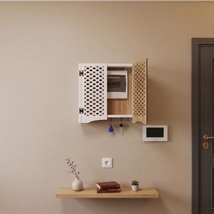 May include: A white wall-mounted cabinet with a lattice-patterned door and a light wood interior, designed to conceal an electrical breaker box. The unit features a row of hooks underneath for hanging keys and is mounted above a floating wood shelf.