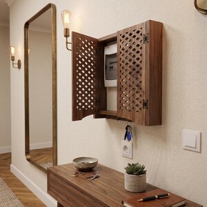 May include: A wall-mounted wooden cabinet with two lattice-patterned doors that open to reveal an electrical breaker box. The cabinet features small hooks on the bottom edge for hanging keys. The wood has a rich brown grain and finish.
