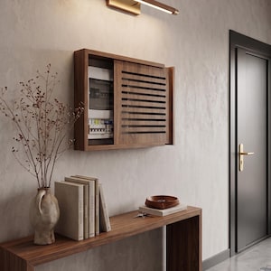 Op de afbeelding: Een rechthoekige houten wandafdekking voor een meterkast met horizontale lamellen en een schuifdeurmechanisme. De unit is aan de muur bevestigd boven een houten consoletafel, waarop een keramische vaas met gedroogde takken en boeken staat.