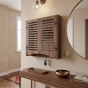 May include: A wall-mounted wooden key cabinet featuring two louvered doors with decorative metal hinges. The cabinet includes a row of metal hooks along the bottom edge for hanging keys, with a set of keys currently suspended from one hook.