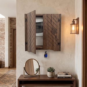 May include: A wall-mounted dark wood cabinet with diagonal slats on the doors, featuring a set of keys hanging from the bottom. Below sits a dark wood console table with a round gold-framed mirror, a small potted succulent, and two stacked books.