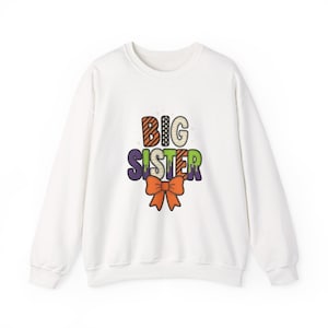 May include: A white long-sleeve crewneck sweatshirt featuring the text BIG SISTER in a multi-coloured, patterned font with a dripping effect. Below the text is a large orange bow graphic. The design includes small star accents.
