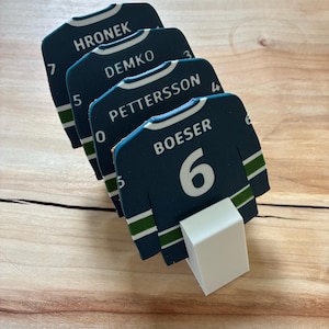 Könnte beinhalten: Ein Set aus vier dunkelblauen Untersetzern in Form von Eishockeytrikots mit weißem Text und Zahlen. Die Namen HRONEK, DEMKO, PETTERSSON und BOESER sind zusammen mit den jeweiligen Trikotnummern aufgedruckt. Ein weißer Kunststoffständer hält die Untersetzer.