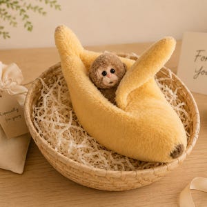 May include: A plush brown monkey toy nestled inside a soft yellow banana-shaped bed. The banana bed has a fuzzy texture with a dark brown tip. The monkey has dark eyes and a light-colored face. The toy is placed in a round woven basket filled with paper filler.