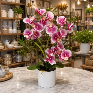 May include: A potted artificial orchid plant featuring vibrant pink and white petals with yellow centers, green stems, and unopened buds. The plant is housed in a simple, cylindrical white ceramic pot, resting on a white marble surface.