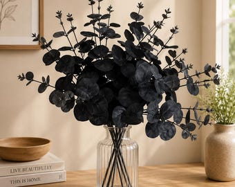 May include: A bouquet of artificial black eucalyptus stems with round leaves arranged in a clear, vertically ribbed glass vase. The stems are thin and dark, supporting multiple branches of foliage that spread outward in a full, rounded shape.