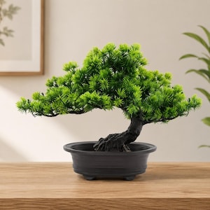 May include: A small artificial bonsai tree with vibrant green needle-like foliage and a textured dark brown trunk. The tree is planted in a simple, matte black oval-shaped plastic pot with a wide rim and a low profile base.