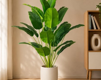 May include: A tall artificial bird of paradise plant with large, vibrant green leaves and slender stems, potted in a smooth, cylindrical off-white ceramic planter. The plant features multiple broad, oval-shaped foliage arranged in a dense, upright cluster.