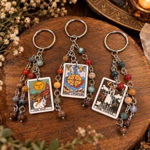 May include: Three metal keychains featuring rectangular tarot card charms labeled The Sun, Wheel of Fortune, and The Tower. Each keychain includes a silver split ring and two dangling chains adorned with various multicolored stone beads.