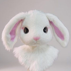 Puede incluir: Una máscara de cabeza de conejo de peluche blanco con pelaje largo y suave, ojos grandes y oscuros, y detalles rosados en el interior de las orejas y la nariz. La máscara está montada sobre un soporte de madera, mostrando un diseño detallado.