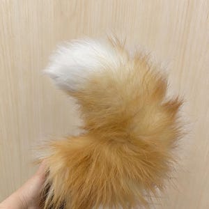 May include: A fluffy, curved faux fur tail featuring a gradient of golden-brown and orange tones that transition into a bright white tip. The long, soft fibers create a voluminous, textured appearance, mimicking the shape of a fox tail.