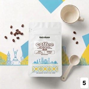 May include: A white coffee bag labeled Dobrakava Coffee Company with the text coffee, decaf, notes of caramel, dried fruit, vanilla, citrus, and geography South America Columbia. Includes a white ceramic mug, a white scoop, and scattered coffee beans.