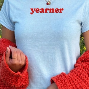 Op de afbeelding: Een lichtblauw T-shirt met korte mouwen en het woord yearner in rode kleine letters op de borst. Het shirt wordt gedragen onder een fel oranje, getextureerd vest en een gouden ketting met een kleine hanger is zichtbaar.