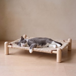 May include: A rectangular pet bed with a light-colored wooden frame and four rounded legs. The sleeping surface is made of a woven, textured tan material stretched across the frame. A grey and white cat is sleeping on the bed.