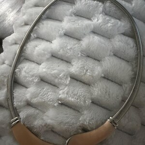 May include: A silver-toned metal snake chain necklace featuring a smooth, curved, beige-colored accent piece at the center. The necklace has a polished finish and a secure clasp mechanism at the top, creating a continuous circular shape.