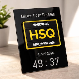 May include: A black rectangular plaque featuring the text Mixtes Open Doubles, 11 Avril 2026, and 49:37. Centered is a black and yellow patch with the text VAUDREUIL, HSQ, and DEMI_HYROX 2026. The plaque has a glossy finish and stands upright.