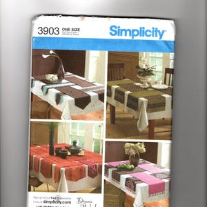 May include: Simplicity sewing pattern 3903 for table runners and placemats. The pattern shows four different table setting designs with various color schemes, including brown, green, red, and pink. The pattern is one size.