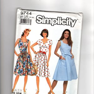 Op de afbeelding: Een Simplicity 9744 naaipatroon-envelop voor maten U5 (16-24). De omslag toont illustraties van drie jurken: één met een blauwe bloemenprint, één met rode stippen en één effen lichtblauwe jurk, allemaal met strikceintuur en zakken.