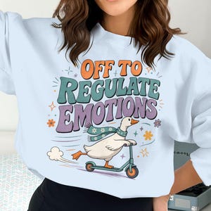 Op de afbeelding: Een lichtblauw sweatshirt met lange mouwen en een afbeelding van een witte eend met een blauwgroene bloemrijke sjaal op een blauwgroene step. De tekst "OFF TO REGULATE EMOTIONS" staat in oranje, blauwgroene en paarse letters, omringd door sterren en bloemen.