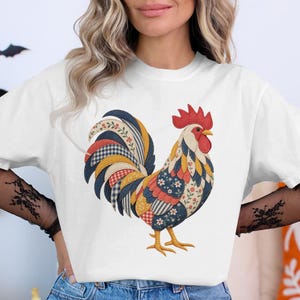 May include: A white short-sleeve t-shirt featuring a large, colorful graphic of a rooster. The rooster is designed with a patchwork-style pattern, incorporating various floral, gingham, and striped prints in shades of red, blue, yellow, and white.
